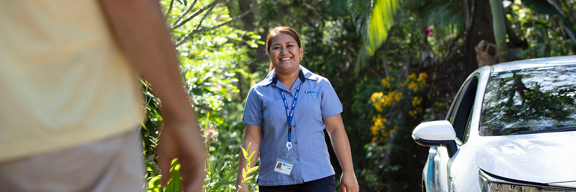 A smiling Blue Care employee stands beside her car as she greets a home care customer.