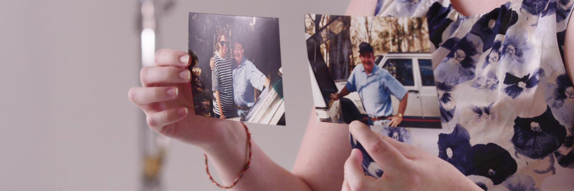 Hands hold family photos of a young woman and her dad standing by a car in the bush.