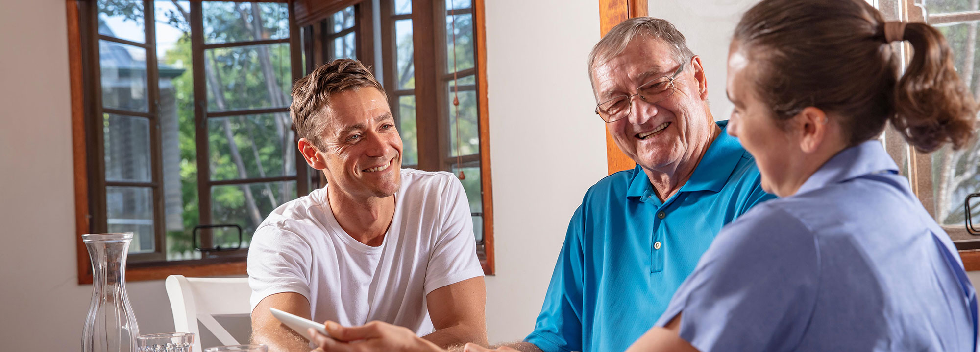 Elderly father and son in living room smiling at Blue Care worker