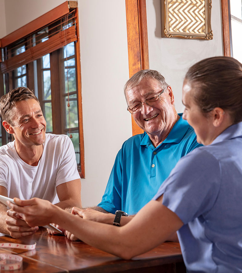 Elderly father and son in living room smiling at Blue Care worker