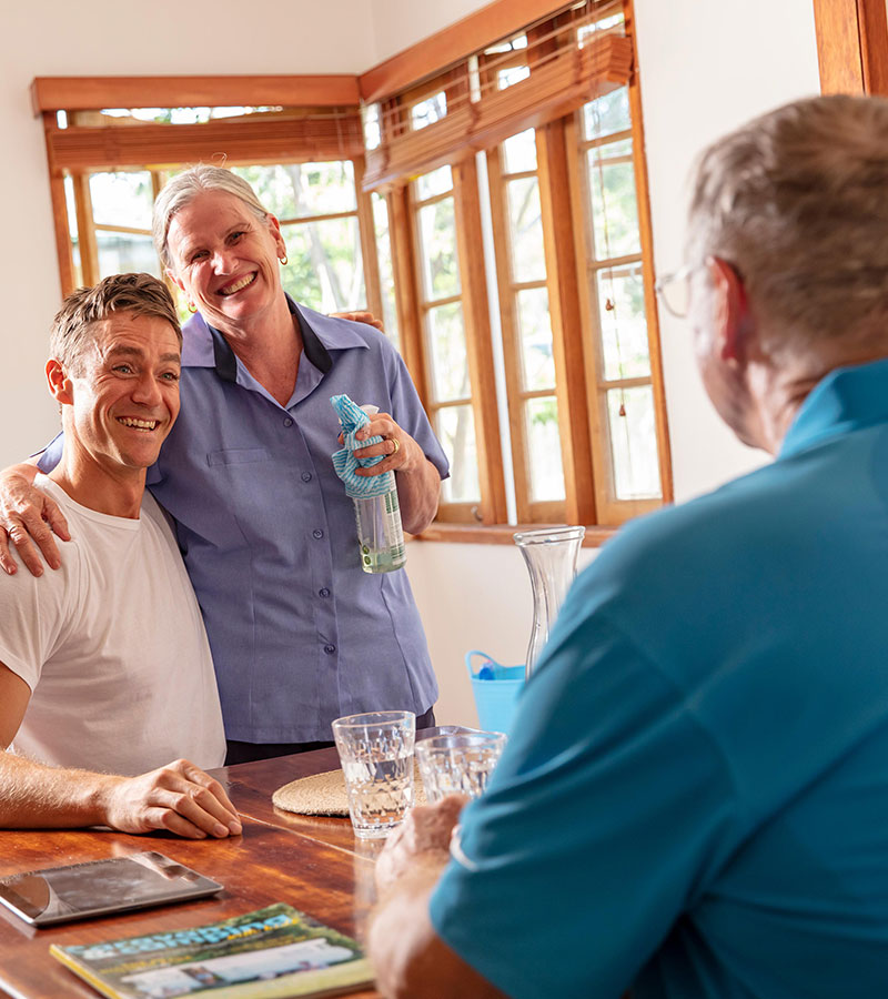 Elderly father and son in living room with Blue Care carer with arm around son