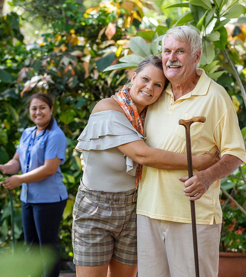 Elderly father and daughter hugging in backyard with Blue Care carer in background watering garden