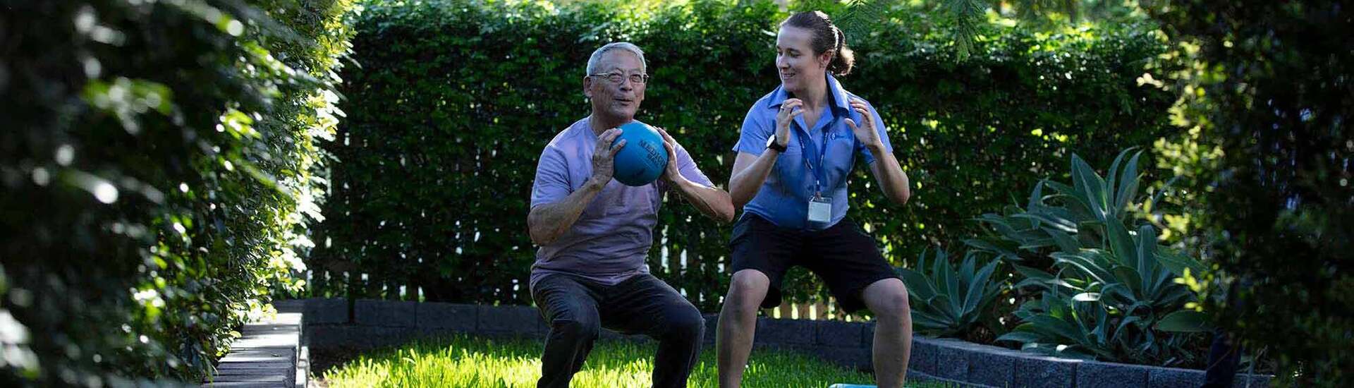 An older man exercising with a medicine ball under the guidance of a Blue Care employee