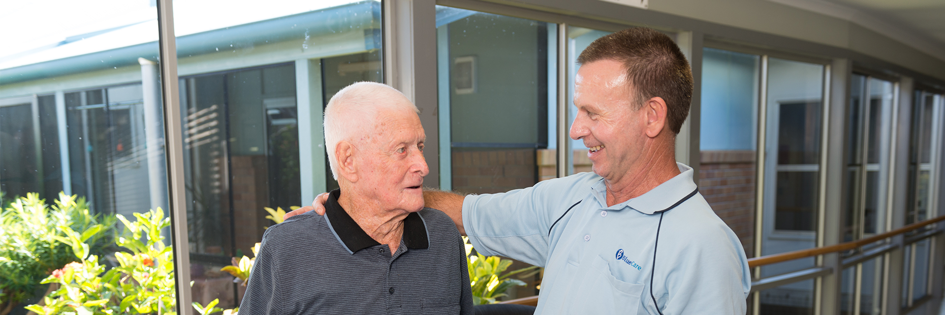 A Blue Care staff member puts his arm around an older gentleman in a common area of Blue Care Bundaberg Riverlea Aged Care Facility.