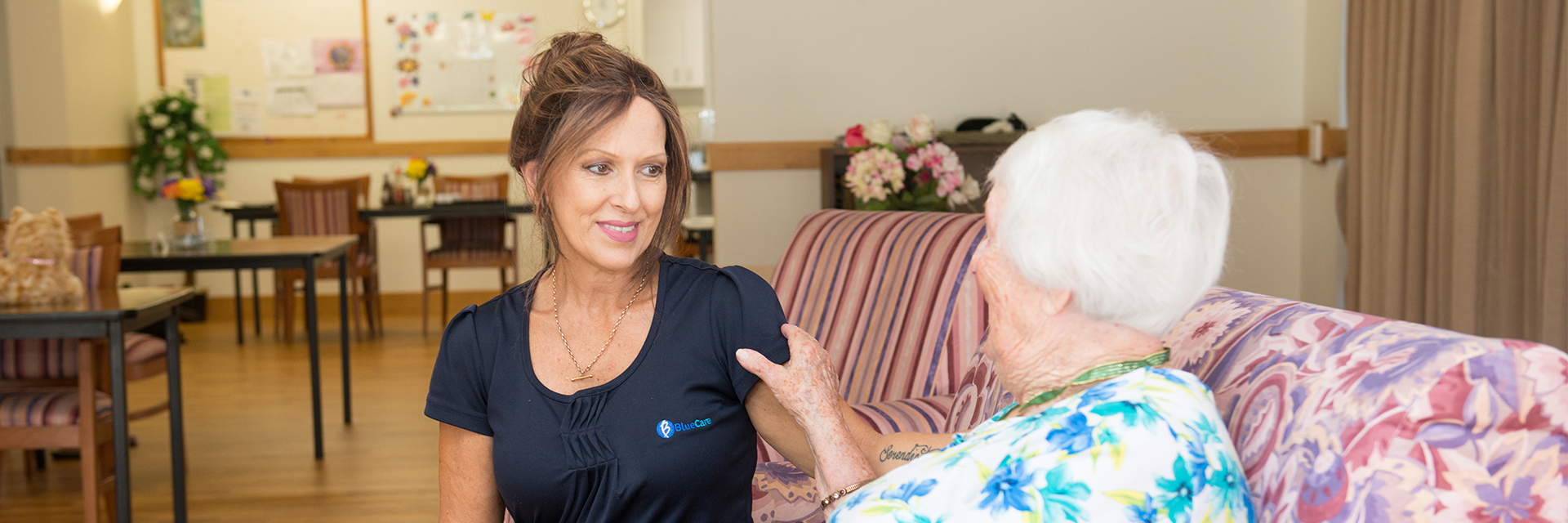 A woman who works at Blue Care Flinders View Nowlanvil Aged Care Facility sits on the couch with a white-haired lady.