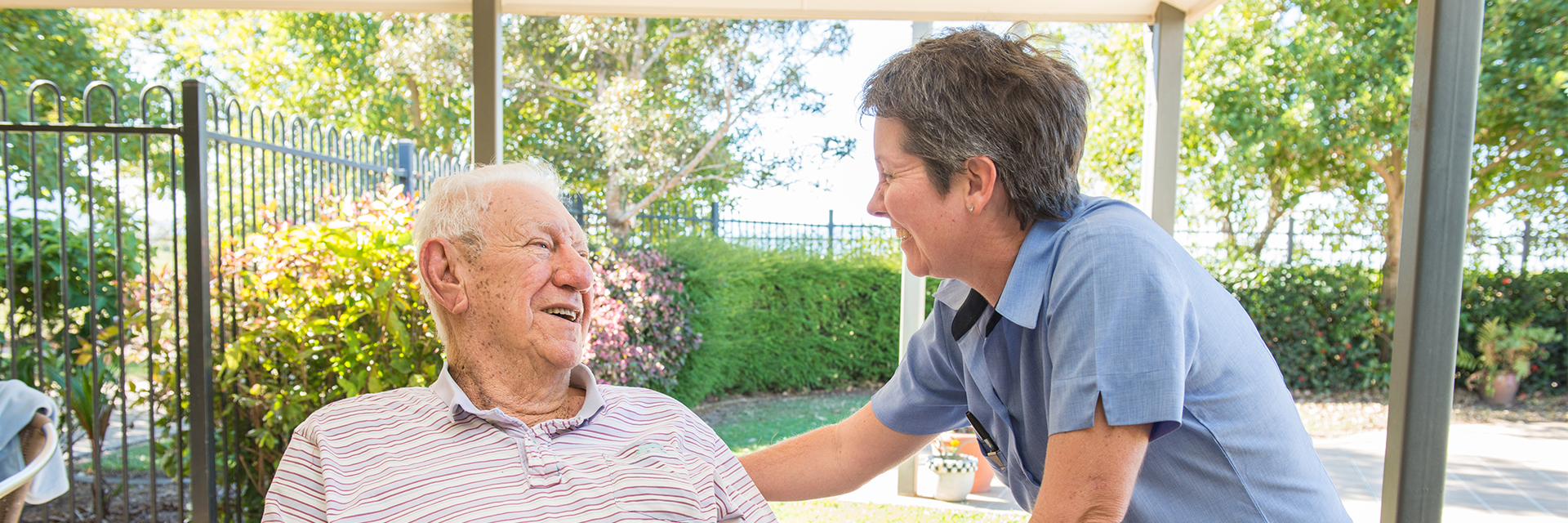 In a shady outdoor area, an older gentleman in a wheelchair has a laugh with a woman who works at Blue Care Ingham Bluehaven Aged Care Facility.