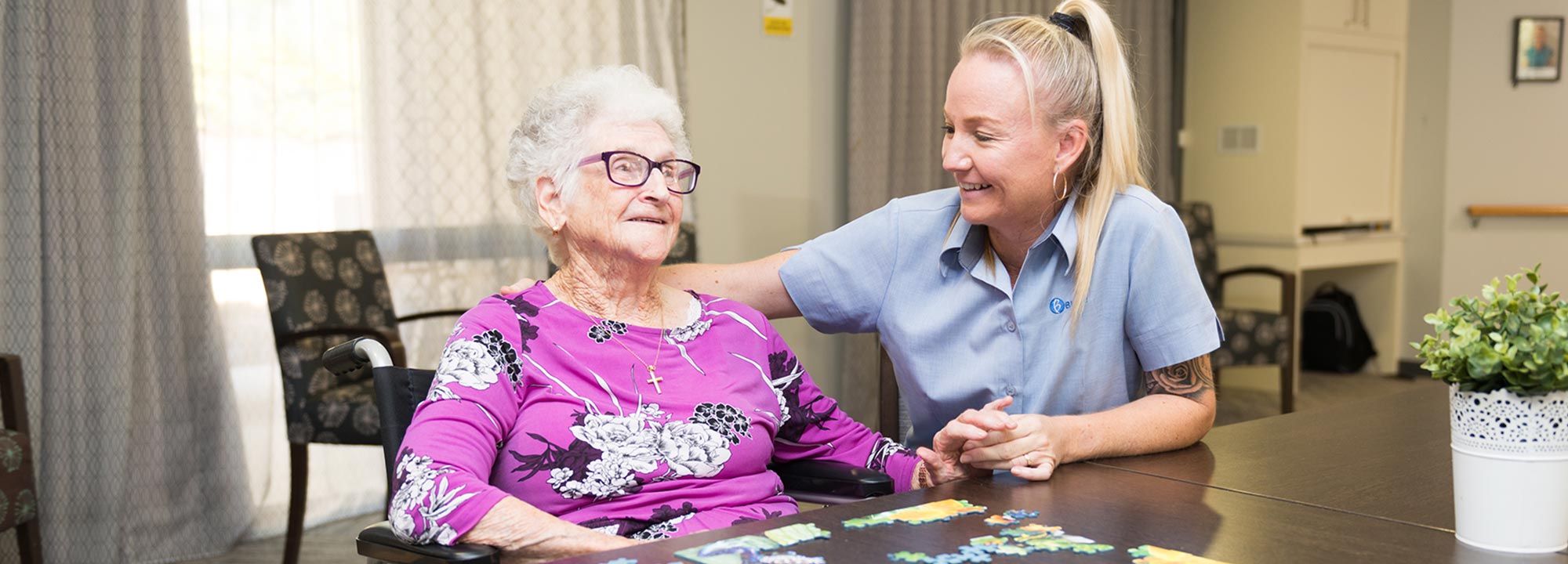 A smiling Blue Care employee with her hand on the shoulder of a smiling older lady in a common area