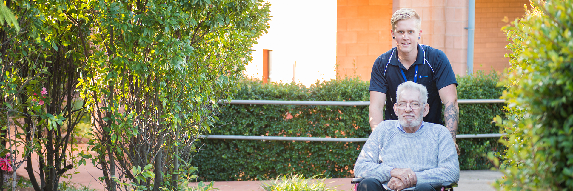 A white-haired man in a wheelchair takes a garden path with the help of a young man who works at Blue Care Toowoomba Aged Care Facility.