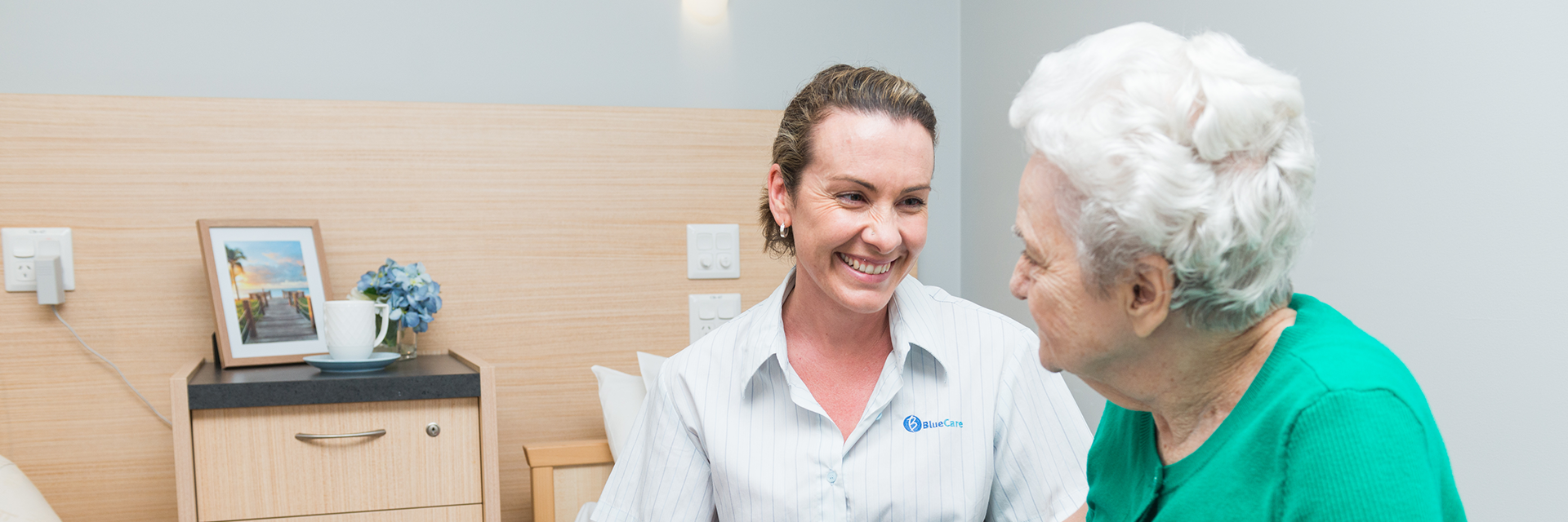 A smiling Blue Care employee looking at an older lady in her room