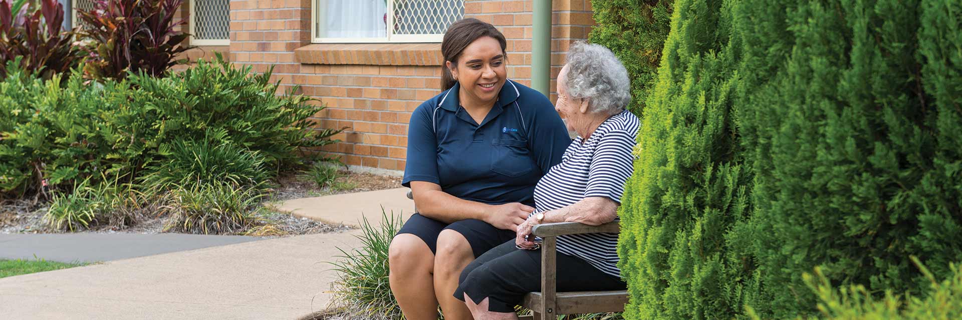 A smiling Blue Care employee holding hands with a smiling older lady in a garden
