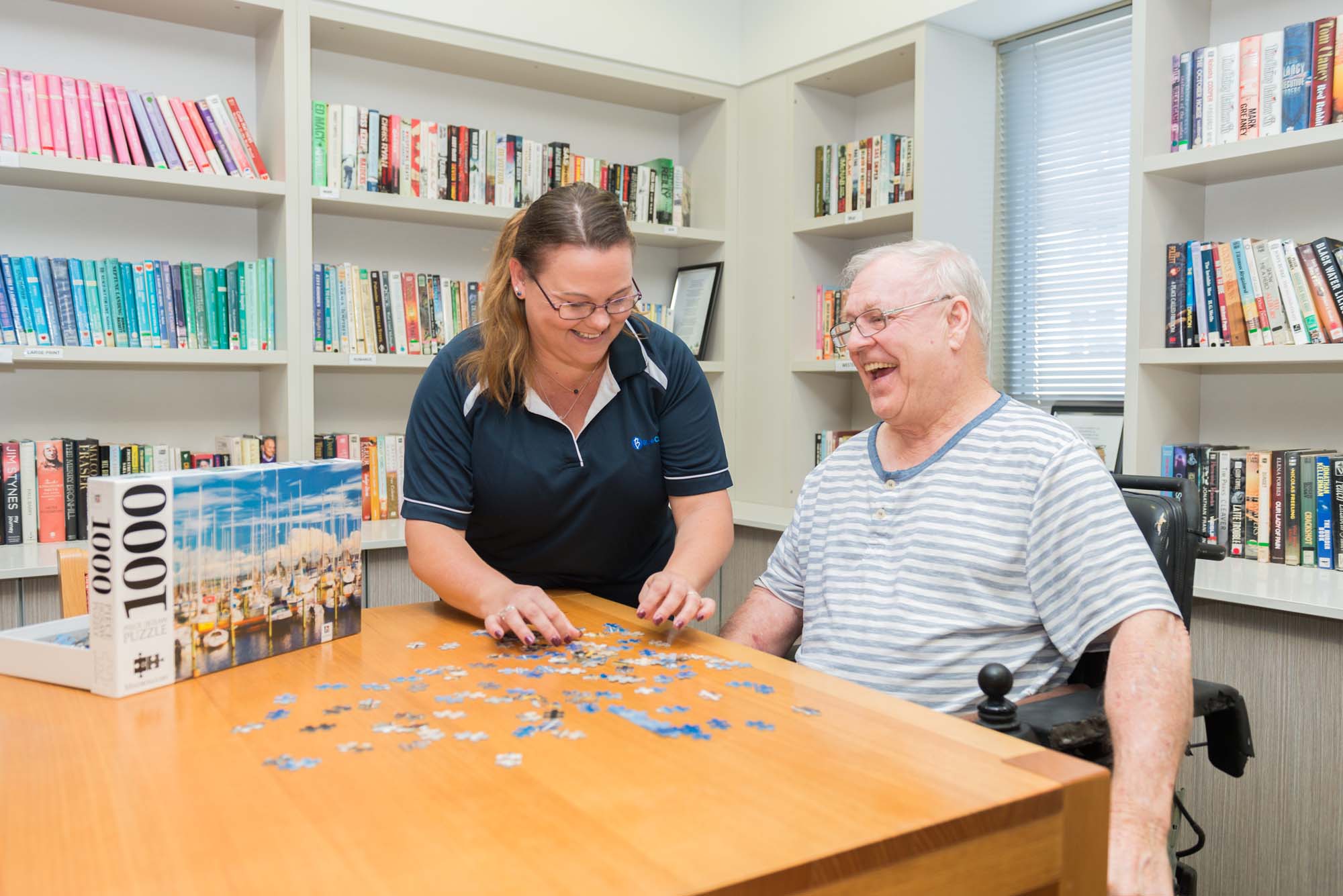 A Blue Care employee completes a jigsaw puzzle with a laughing resident in a common area of Blue Care Carina.