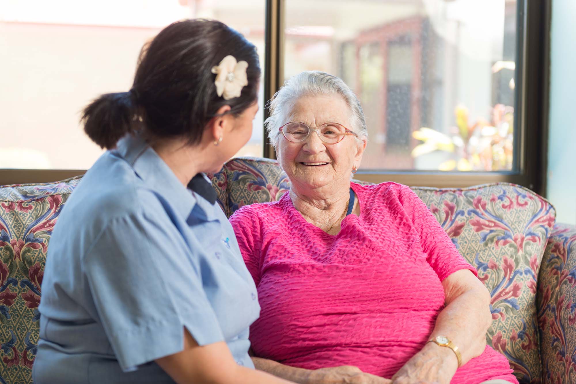 A white-haired lady in a pink top sits on a couch and smiles at an employee at Blue Care Masters Lodge.