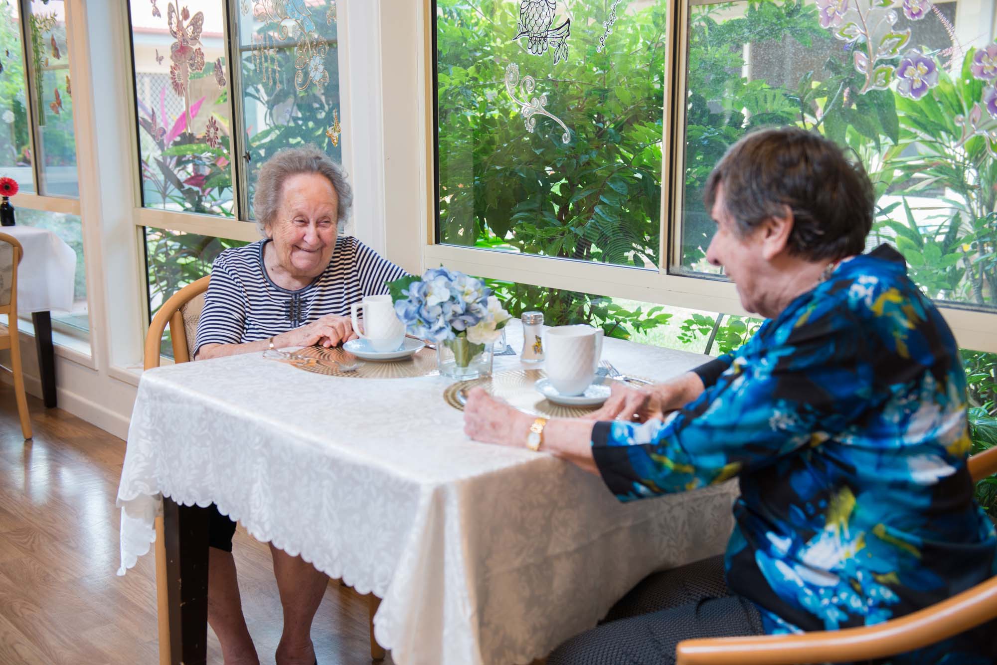 Two residents enjoy a laugh over tea in the dining room of Blue Care Wynnum, with views of the lovely garden.