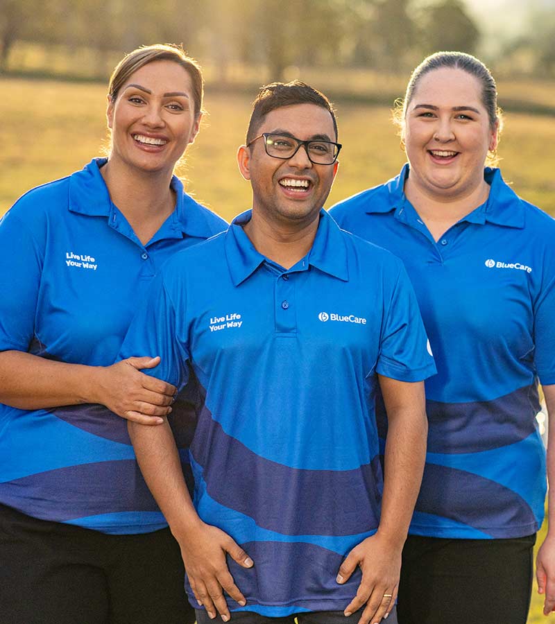 Three BlueCare staff standing together in a field in front of a fleet of BlueCare vehicles, smiling at camera