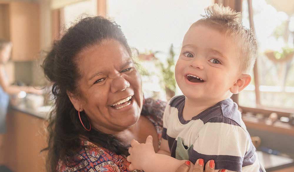 elderly indigenous woman holding an infant child as they both laugh looking into the camera