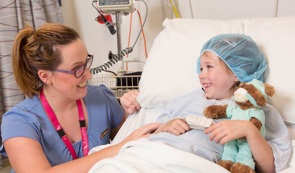 young girl laying in a hospital bed with a hairnet on holding a teddy bear smiling as a female nurse comforts her