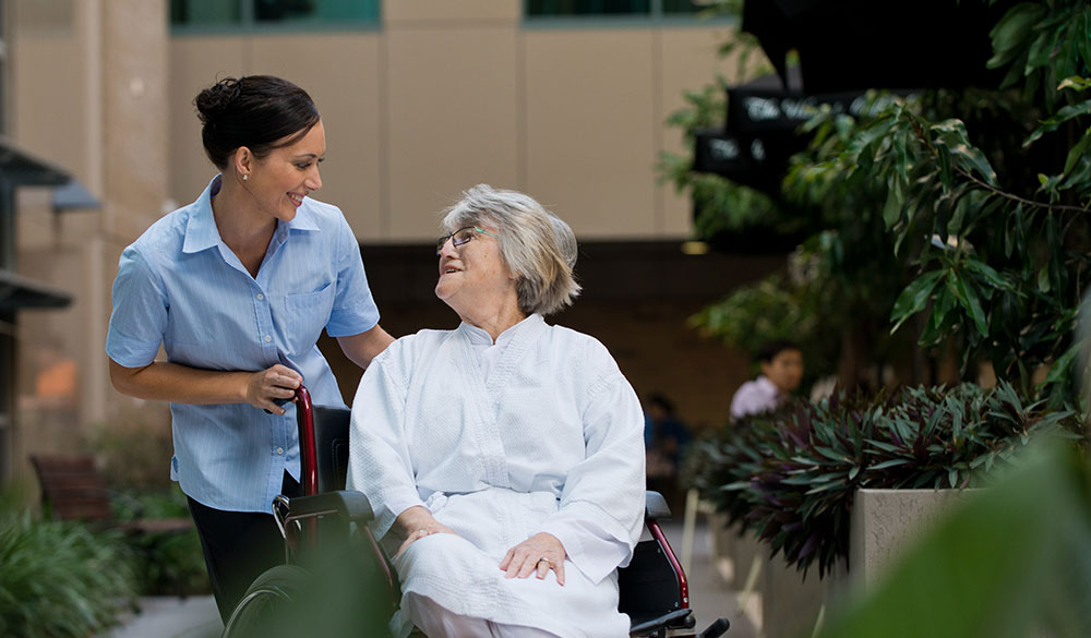 elderly female patient sitting in a wheelchair outdoors looking up towards the female staff member standing beside her