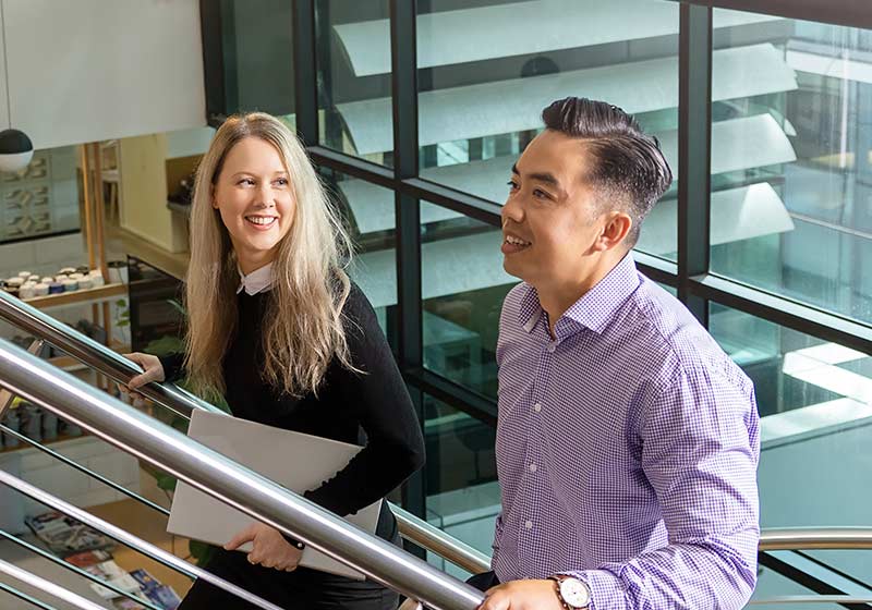 Two UnitingCare staff members climb the stairs in UnitingCare's head office, with city buildings visible through the glass walls behind them.
