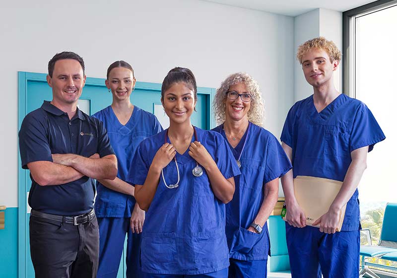 Five hospital workers, some wearing scrubs, stand in a blue hospital corridor, smiling.