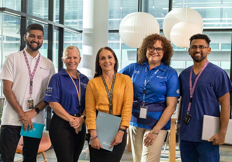 Five UnitingCare staff members, from across the organisation stand in head office, with city buildings visible through the glass walls.