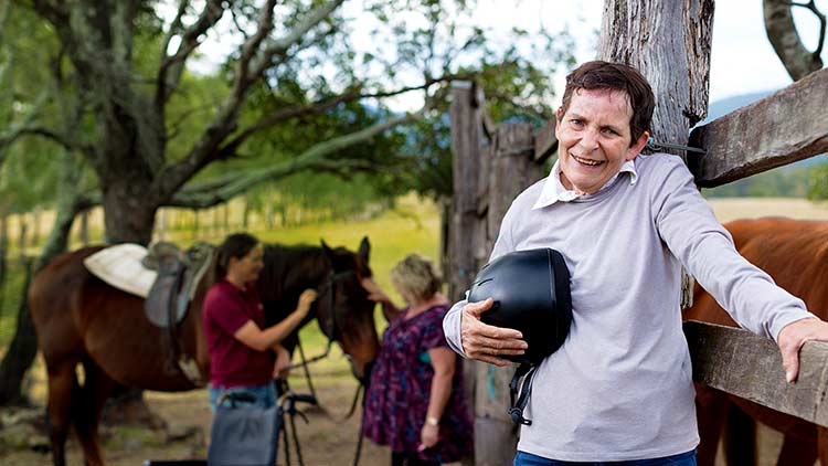 disabled man leaning against fence outdoors with staff attending a horse in the background