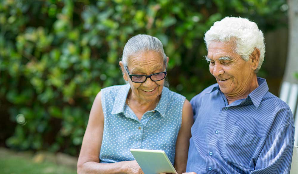 elderly couple sitting outside smiling while looking at ipad