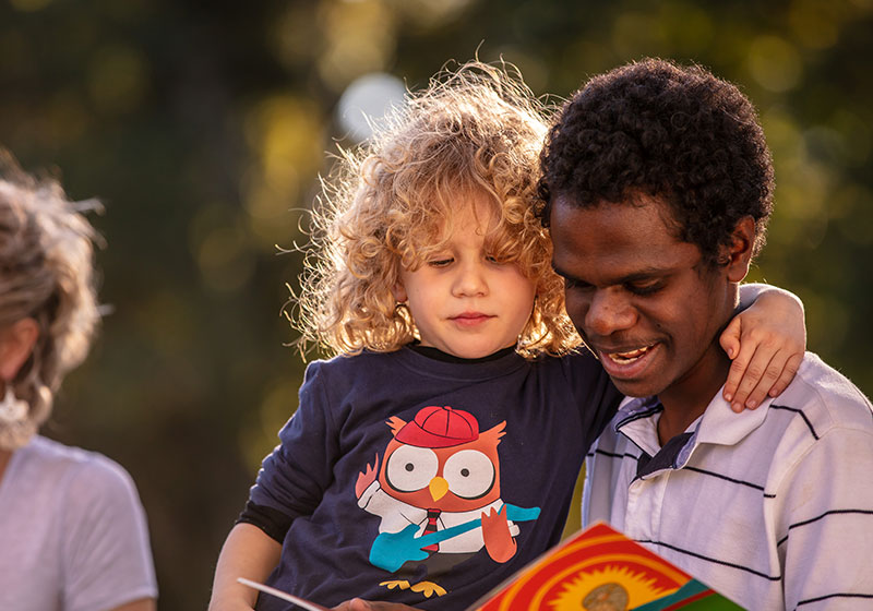 indigenous carer holding curly headed child reading outside