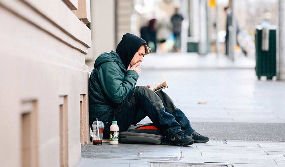 homeless man sitting on city street smoking and reading