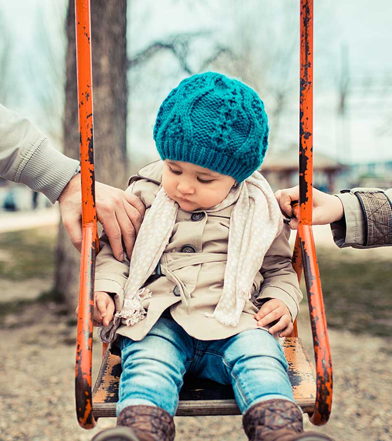 A young child in a beanie plays on a swing with a parent on either side.