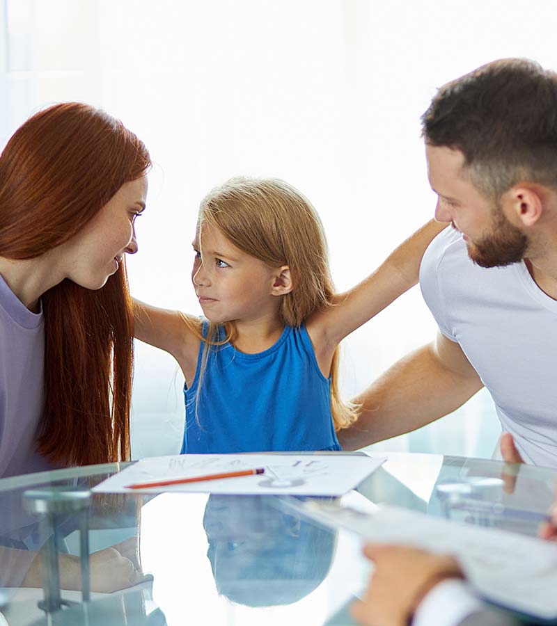 A young girl stands between her mother and father, who are sitting at a table with a mediator.