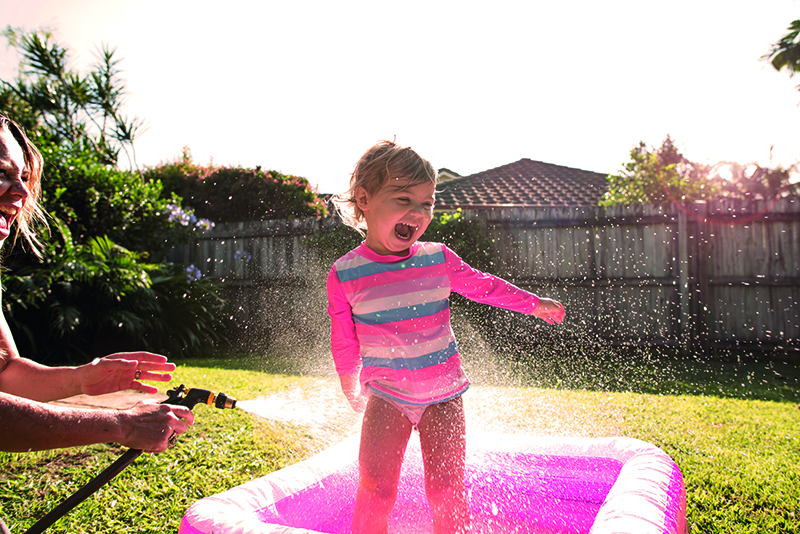 UnitingCare Early Childhood Approach A young girl in a blow up kids pool being sprayed with a hose be her mum in her back yard