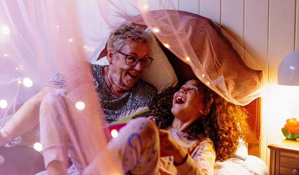 Grandmother and granddaughter laughing in bed surrounded by fairy lights