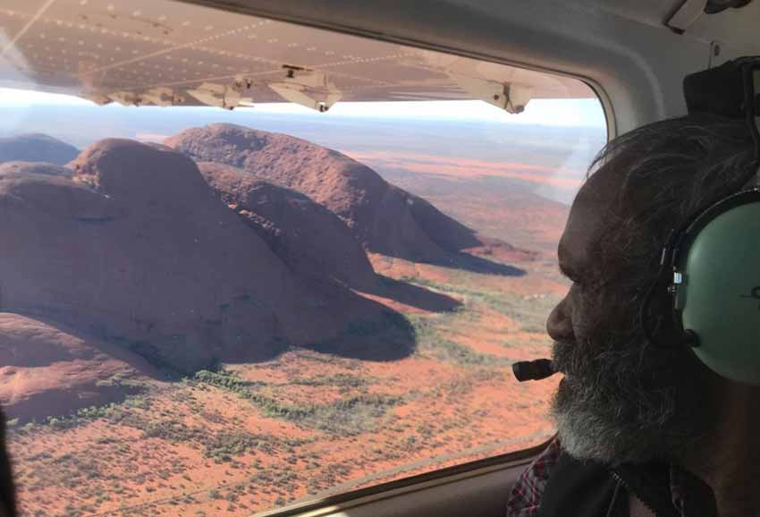 An Aboriginal Elder wearing headphones looks out the window of an aeroplane on to a stunning view of Kata Tjuta.