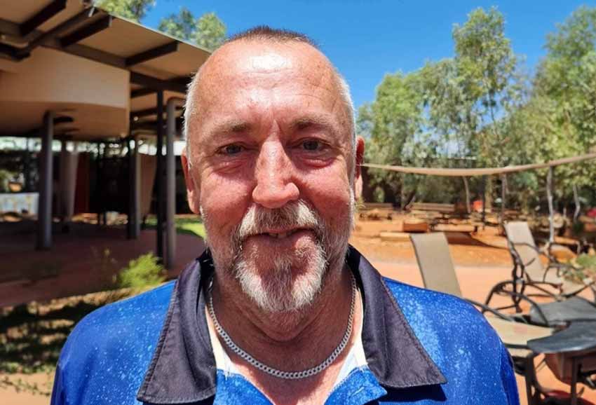 ARRCS Activities Officer Todd Rayner wears a blue ARRCS polo shirt with the red earth of the Nganampa Ngura Mutitjulu-nya Flexible Aged Care facility in the background.