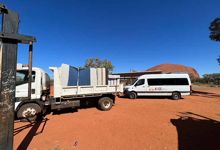 A truck loaded with mattresses and an ARRCS van stand on the red dirt before Uluru, ready to deliver secondhand furniture to the community of Mutitjulu. 