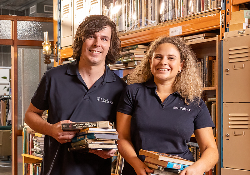 A young man and woman, wearing Lifeline T-shirts, stand in a Lifeline Shop surrounded by floor-to-ceiling shelves filled with books.