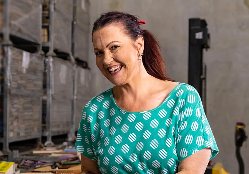 A lady standing in a warehouse holding books beside large containers filled with books, smiling at camera