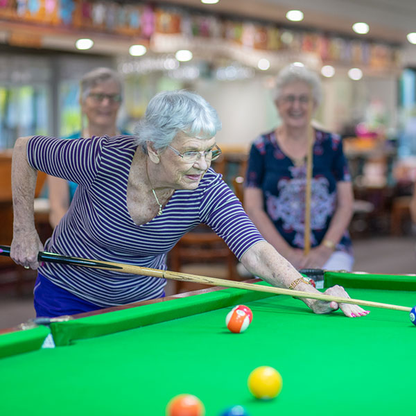 An elderly lady playing billiards and people watching in the background