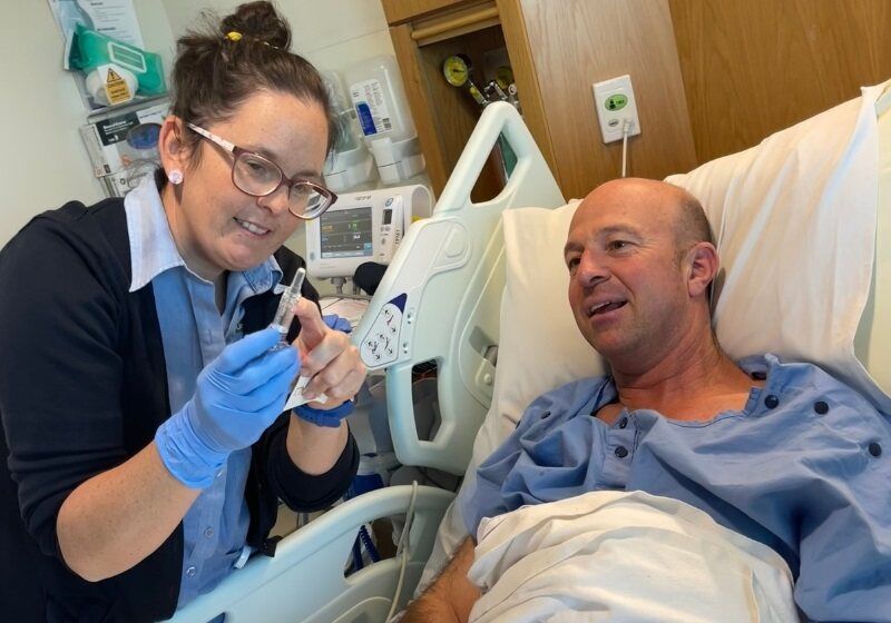 Patient Andrew Parry lies in a hospital bed, talking to a smiling nurse.