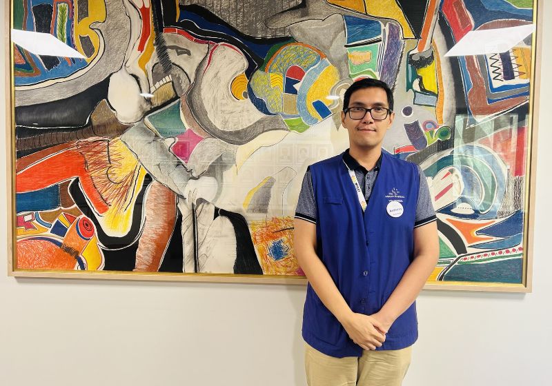 Anthony Kusoy, wearing a purple volunteer vest, stands in front of a colourful artwork at The Wesley Hospital.