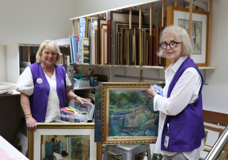 Wesley Hospital volunteers Caroline McCormack and Kathy Praine, wearing purple volunteer vests, stand surrounded by artworks. 