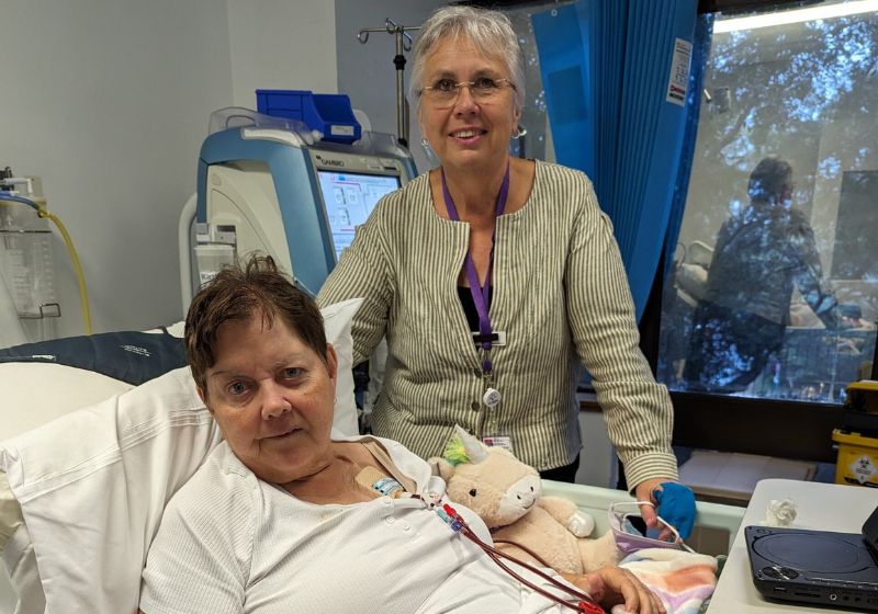 Volunteer Compassionate Companion Lyndie Mitchell stands beside a patient, Vicky, who is lying in bed. 