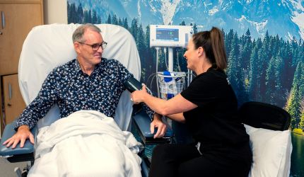 A female nurse takes the blood pressure of a male patient sitting in a hospital chair with a blanket on his lap.
