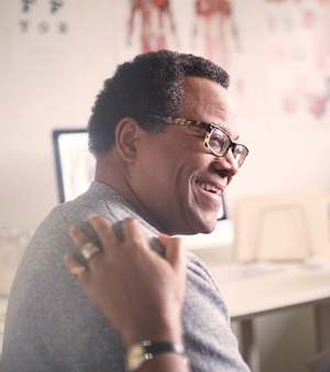 An elderly gentlemen smiling at a person with their hand on his back off camera