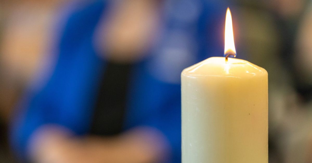 A close up of a candle with a person with folded hands in the background