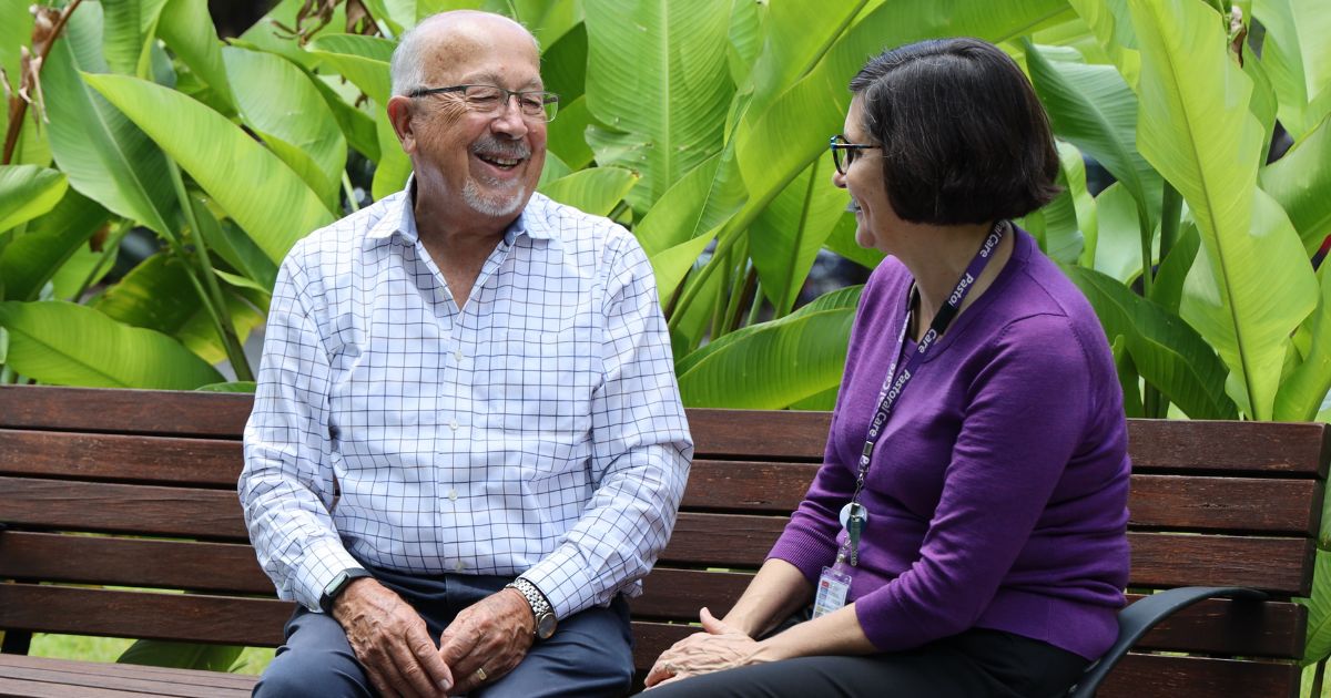 A man in a white checked shirt and a woman in a blue jumper sit on a park bench backed by greenery.